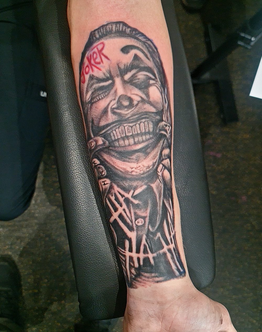 Black and grey tattoo of the Joker with red lettering on a client's forearm