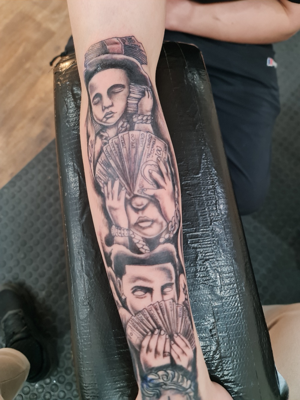 Black and grey realism sleeve tattoo of three faces with cash motifs on a forearm